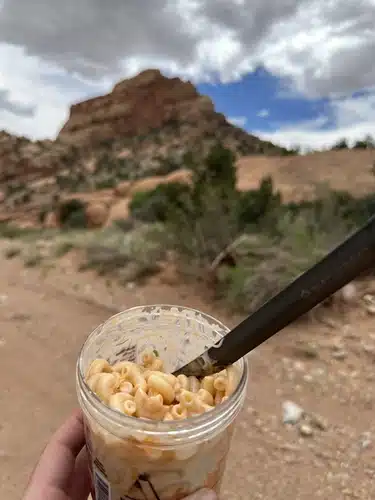 Hand holding a Talenti jar of cold soak buffalo pasta salad with a spoon on a desert trail, red rock formations in the background.