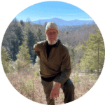 Male hiker wearing a beanie and outdoor gear, posing with a scenic mountain landscape in the background on a clear, sunny day.
