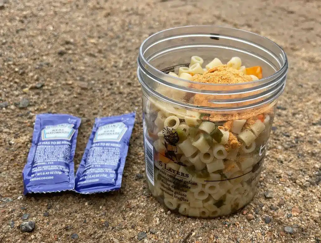 Talenti jar filled with dry pasta, vegetables, and buffalo seasoning mix for backpacking pasta salad, with two mayonnaise packets placed beside it on the ground.