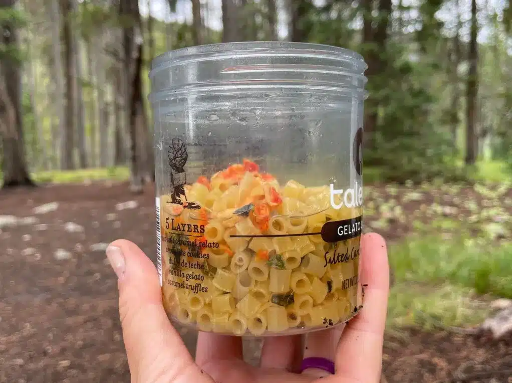 Hand holding a Talenti jar filled with dry pasta, vegetables, and seasonings for buffalo pasta salad, ready to cold soak on a backpacking trip in the forest.