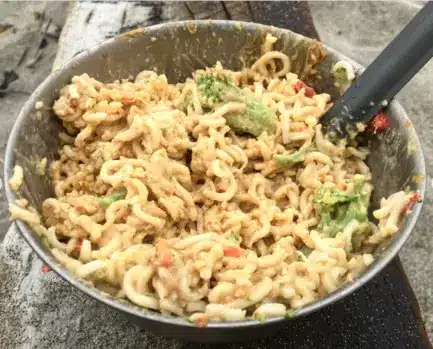 Prepared cold-soak pasta salad in a metal bowl with a spoon, after being transferred from an overfilled container.