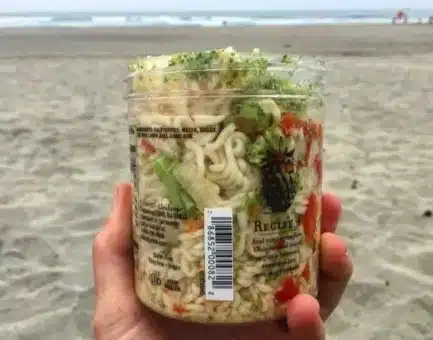 Cold-soaked backpacking pasta salad in a repurposed Talenti jar, shown on a beach with a view of the ocean.