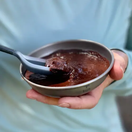 Backpacker holding a camping bowl of prepared Brownie Batter Hummus dessert with a spoonful lifted, ready to eat