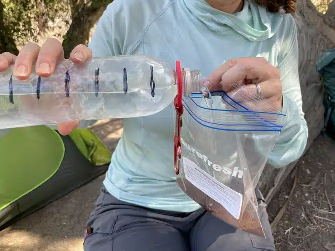 Backpacker pouring water from a marked plastic bottle into a resealable bag with cold soak dessert mix while camping outdoors