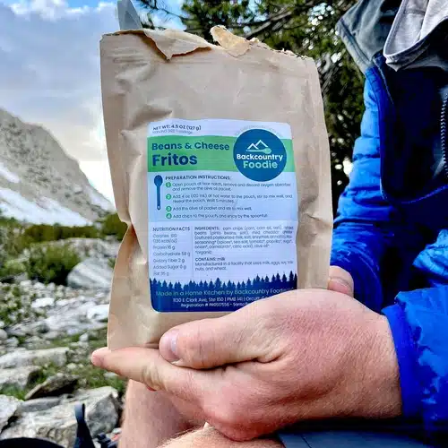 Hiker holding a Backcountry Foodie Beans & Cheese Fritos backpacking meal pouch in a mountainous outdoor setting with trees and rocky terrain.