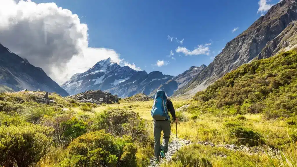 Backpacker with a blue pack hiking through a lush alpine valley surrounded by dramatic mountains under a partly cloudy sky.