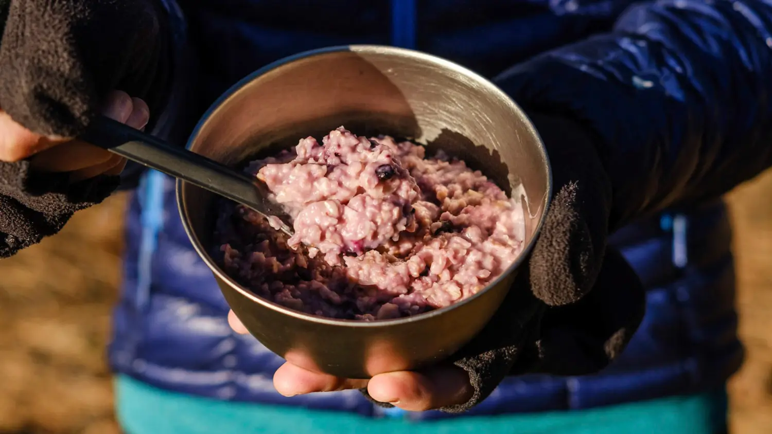 Warm lemon blueberry oatmeal served in a metal camping bowl, held by a hiker wearing gloves on a chilly morning