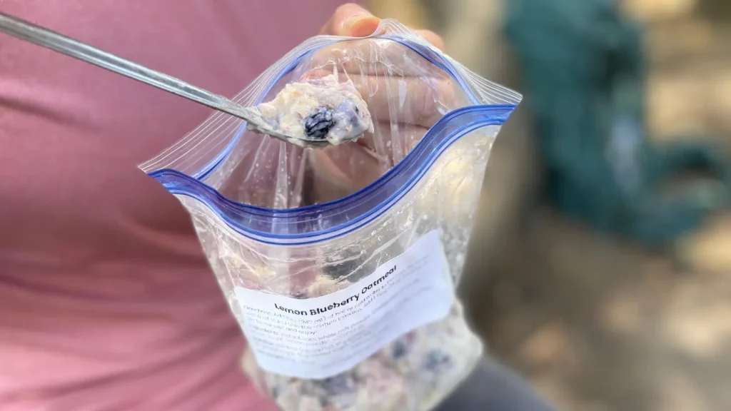 Backpacker eating lemon blueberry oatmeal straight from a labeled freezer bag using a spoon — a quick, high-calorie trail breakfast