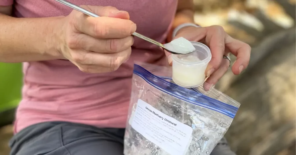 Backpacker scooping coconut oil from a small container to add to lemon blueberry oatmeal in a freezer bag — ultralight trail breakfast prep