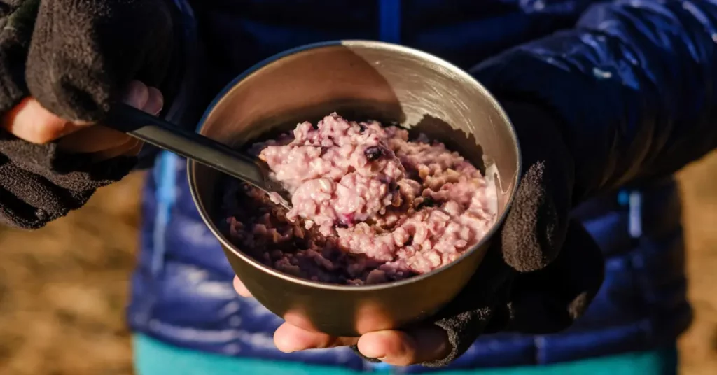 Backpacker holding a warm bowl of lemon blueberry oatmeal, a high-calorie vegan backpacking breakfast, ready to eat on a cold morning