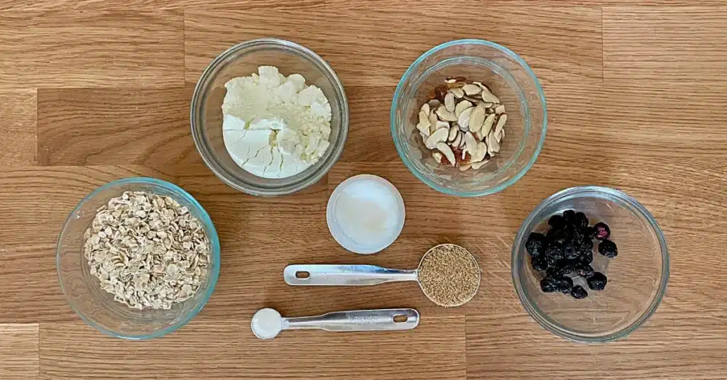Ingredients for lemon blueberry backpacking oatmeal laid out in glass bowls, including oats, milk powder, almonds, coconut oil, brown sugar, and freeze-dried blueberries