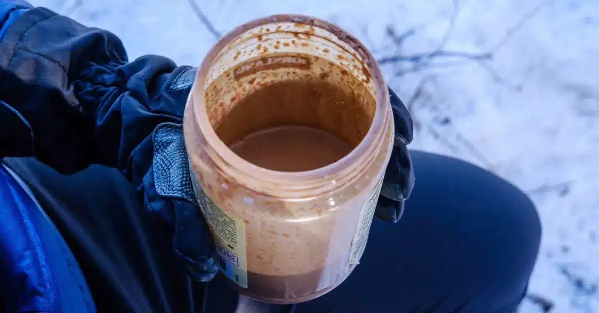 Ultralight backpacking protein shake mixed in an empty peanut butter jar, held by hiker wearing gloves in snowy backcountry