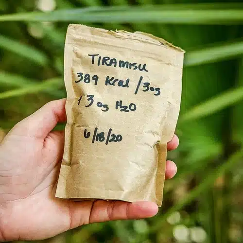 Hand holding a labeled brown paper bag of cold soak tiramisu backpacking dessert with calories, serving size, and water instructions written on it