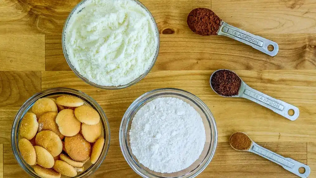 Backpacking tiramisu ingredients in glass bowls and measuring spoons: powdered milk, vanilla pudding mix, vanilla wafers, cocoa powder, espresso powder, and cinnamon