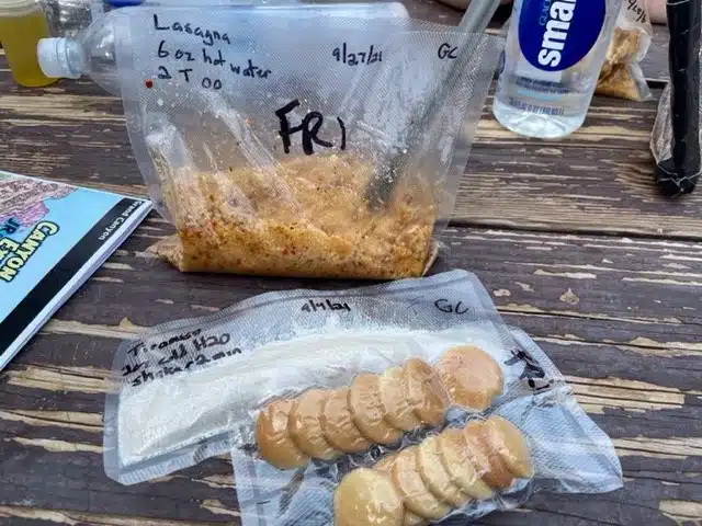Vacuum-sealed backpacking meals on a picnic table, including lasagna with water instructions and a cold soak tiramisu dessert with cookies packed separately