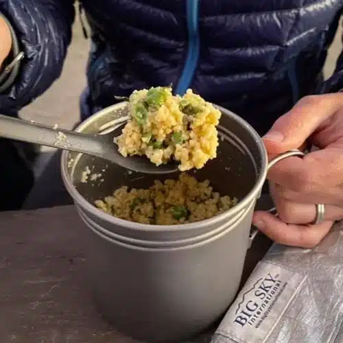 Backpacker holding a titanium mug filled with rehydrated couscous and vegetables, using a long-handled spoon to take a bite; sitting at a wooden table with a Big Sky International insulated pouch nearby.