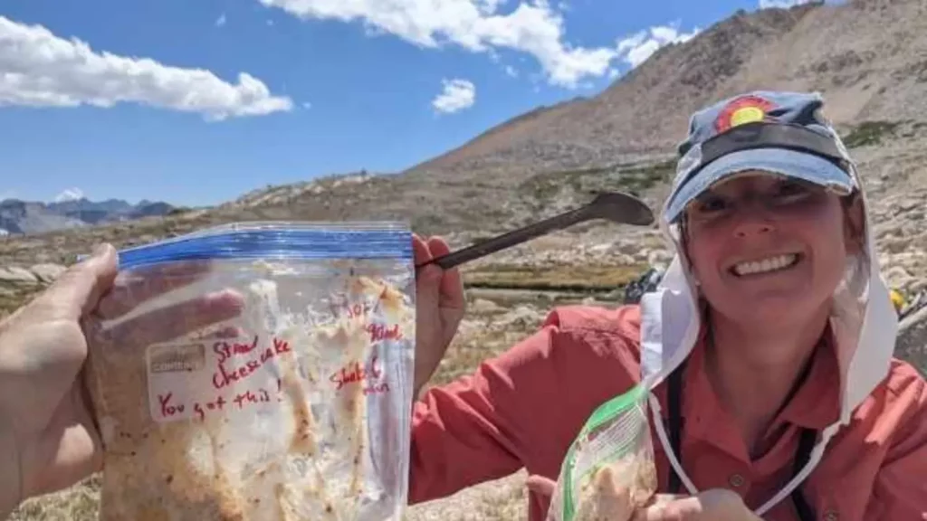 Smiling hiker holding a plastic freezer bag labeled “Strawberry Cheesecake” with a handwritten “You got this!” encouragement, sitting in a high alpine setting with rocky mountains and blue sky in the background.