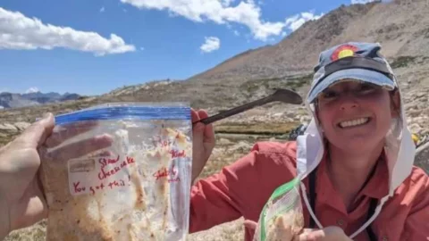 Smiling hiker holding a plastic freezer bag labeled “Strawberry Cheesecake” with a handwritten “You got this!” encouragement, sitting in a high alpine setting with rocky mountains and blue sky in the background.