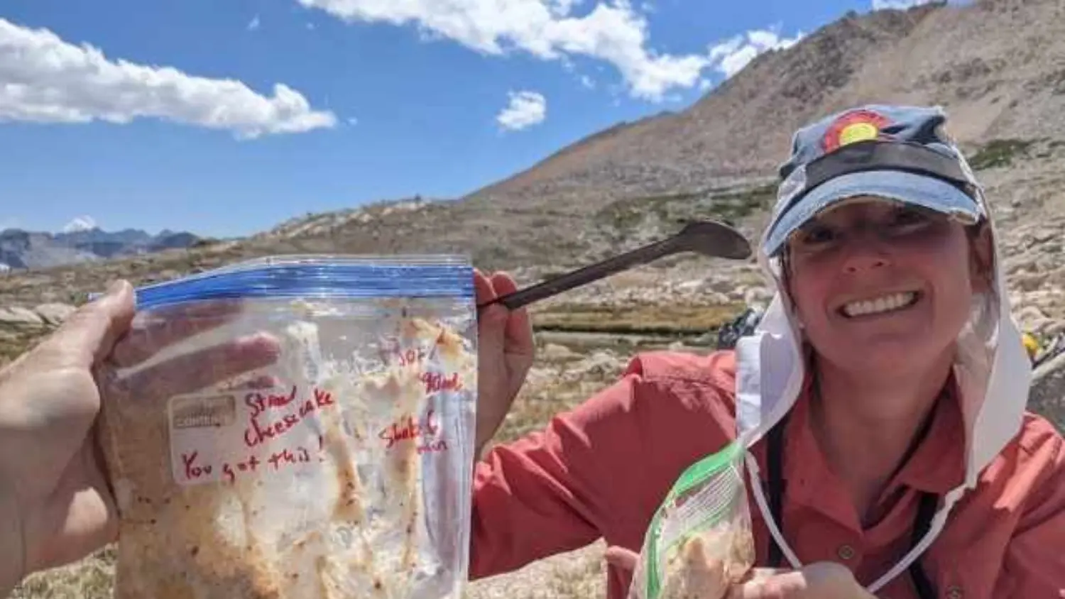 Smiling hiker holding a plastic freezer bag labeled “Strawberry Cheesecake” with a handwritten “You got this!” encouragement, sitting in a high alpine setting with rocky mountains and blue sky in the background.